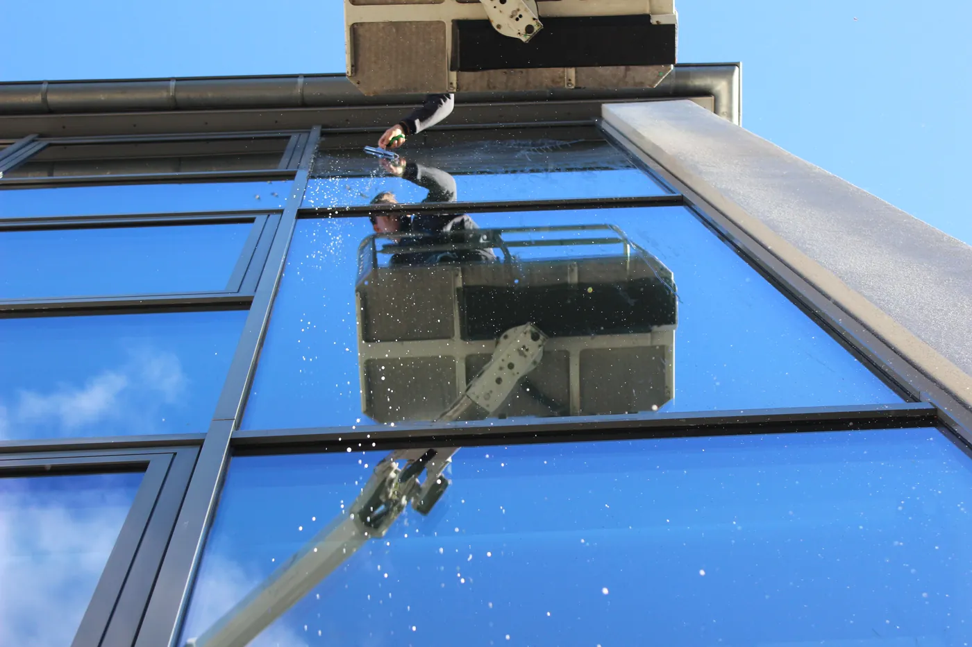 Blick von unten durch eine nasse Glasfassade: Ein Mitarbeiter von G&W reinigt aus dem Korb einer Hebebühne die Scheiben.